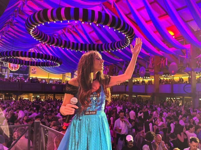 Eine Frau in einem blauen Dirndl steht mit einem Mikrofon auf einer B&uuml;hne und winkt einer gro&szlig;en Menschenmenge in einem festlich beleuchteten Festzelt zu.